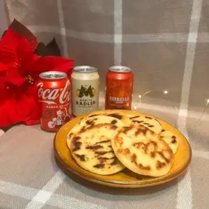 Un plato con Arepas Venezolanas, junto a una coca-cola, estrella damm y estrella damm de limon. Rodeados de una flor de navidad. Experiencias Gastronómicas que debes de probar.